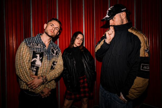 Brighton alt-punk trio SNAYX pose against a red corrugated wall in a press photo for 'Nice Day 4 It'