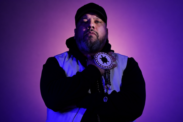 Shub wearing a turtle medallion against a purple backdrop in a press photo for 'Heritage (Part Two).'