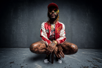 Thundercat crouches in a red and white satin jacket, cap and yellow sunglasses ahead of his 2026 Distracted AF Tour.
