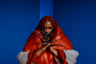 Tierra Whack poses in a bold editorial portrait wearing an oversized red puffer coat with vivid red braids and dramatic black claw accessories, set against a deep blue minimalist background.