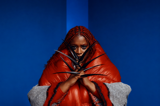 Tierra Whack poses in a bold editorial portrait wearing an oversized red puffer coat with vivid red braids and dramatic black claw accessories, set against a deep blue minimalist background.