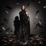 Chelsea Wolfe stands in a dramatic black draped and woven gown under moody lighting, casting a long shadow, in press art for her ninth album 'The Dark.'