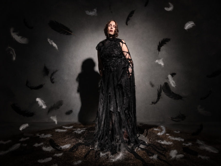 Chelsea Wolfe stands in a dramatic black draped and woven gown under moody lighting, casting a long shadow, in press art for her ninth album 'The Dark.'