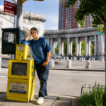 Mike D leans against a yellow newspaper box on a New York City street in a blue sweatshirt and jeans, in press art for his solo debut album 'Thank You.'