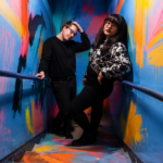 The two members of Sweeping Promises pose in a stairwell covered in bold, colorful abstract paint strokes, both dressed in black, promoting their third album 'You Say I Romanticize.'