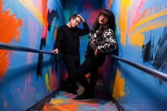 The two members of Sweeping Promises pose in a stairwell covered in bold, colorful abstract paint strokes, both dressed in black, promoting their third album 'You Say I Romanticize.'