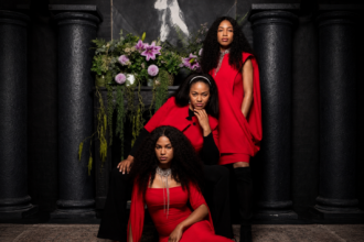 The Womack Sisters, Kucha, Zeimani, and BG Womack, pose in coordinated red-and-black outfits before a dark, columned backdrop with cascading flowers, promoting their self-titled debut album.