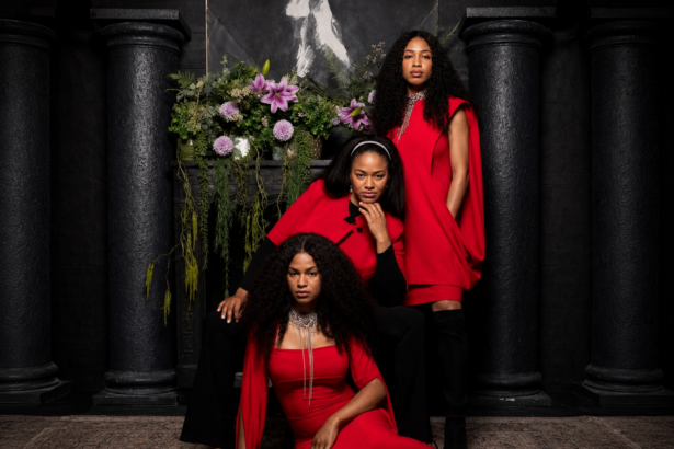 The Womack Sisters, Kucha, Zeimani, and BG Womack, pose in coordinated red-and-black outfits before a dark, columned backdrop with cascading flowers, promoting their self-titled debut album.