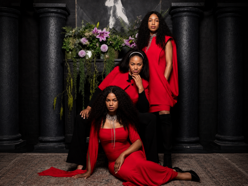 The Womack Sisters, Kucha, Zeimani, and BG Womack, pose in coordinated red-and-black outfits before a dark, columned backdrop with cascading flowers, promoting their self-titled debut album.