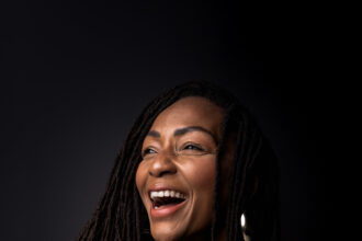 Amana Melome' laughs while holding a small black dog against a dark backdrop, wearing a black outfit and gold jewelry, in press art for her single 'Con C.Alma.'