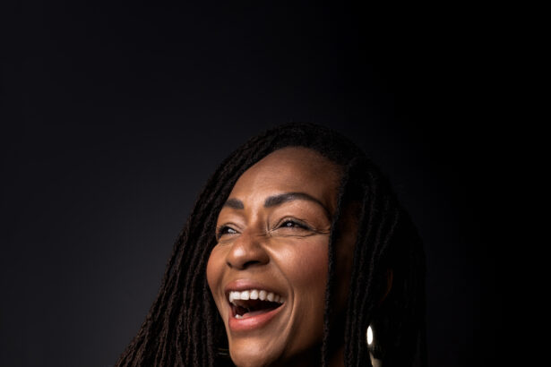 Amana Melome' laughs while holding a small black dog against a dark backdrop, wearing a black outfit and gold jewelry, in press art for her single 'Con C.Alma.'
