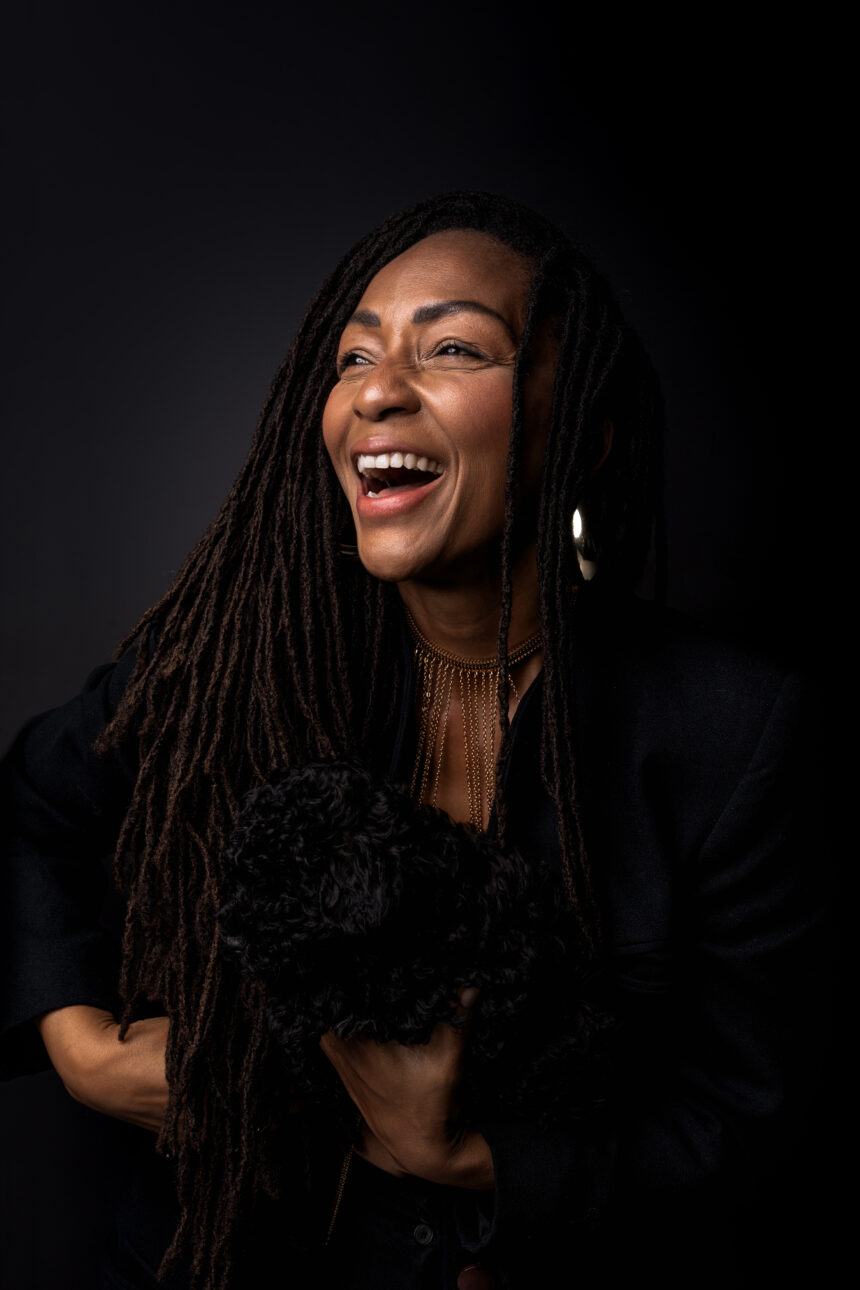 Amana Melome' laughs while holding a small black dog against a dark backdrop, wearing a black outfit and gold jewelry, in press art for her single 'Con C.Alma.'