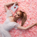 Ariana Grande, whose album 'petal' debuted at No. 1, lies on pink rose petals in a white dress holding a heart-sealed envelope.