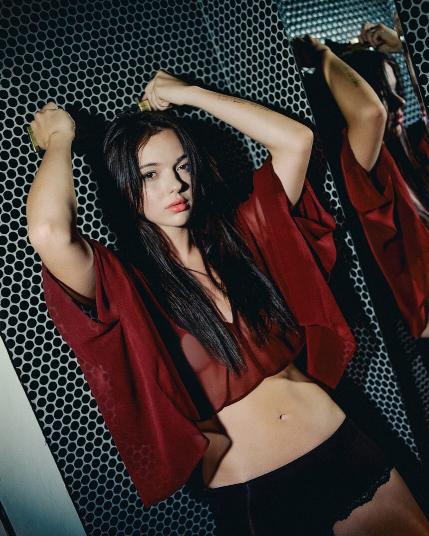 Ava Renn poses against a perforated metal backdrop in a sheer red top, arms raised overhead, in a moody, dramatically lit portrait promoting her single 'You Want Love.'
