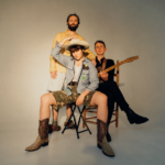 The members of Big Thief pose together in a studio, one seated in a cowboy hat and boots and another holding a guitar, in press art around their triple album 'Horsepower.'