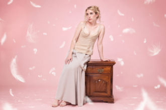Blondshell leans against a wooden nightstand in a cream corset top and long satin skirt against a soft pink backdrop scattered with floating white feathers, promoting her third album 'Violins.'