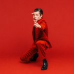 Brandon Flowers, whose single 'Tiger's Blood' precedes his album 'Thrasher,' poses in a red suit against a red backdrop.