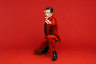 Brandon Flowers, whose single 'Tiger's Blood' precedes his album 'Thrasher,' poses in a red suit against a red backdrop.