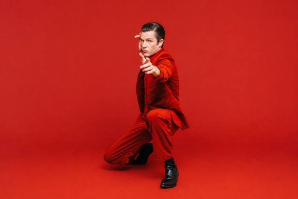 Brandon Flowers, whose single 'Tiger's Blood' precedes his album 'Thrasher,' poses in a red suit against a red backdrop.