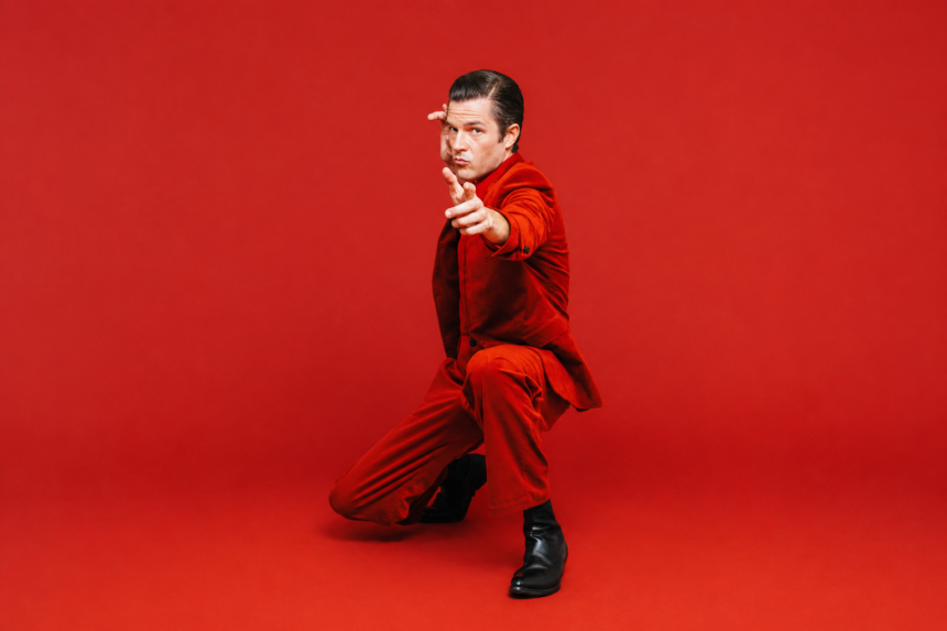 Brandon Flowers, whose single 'Tiger's Blood' precedes his album 'Thrasher,' poses in a red suit against a red backdrop.