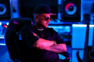ChevoBeatz in a studio, arms crossed under blue and pink lighting.