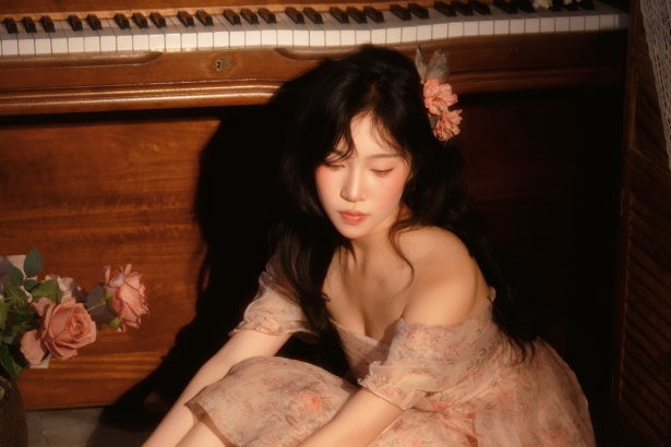 Clarity Liao sits beside a wooden piano in a soft floral off-the-shoulder dress with a flower in her hair, eyes closed in a warm, intimate setting with pink roses, promoting her single 'Have You.'