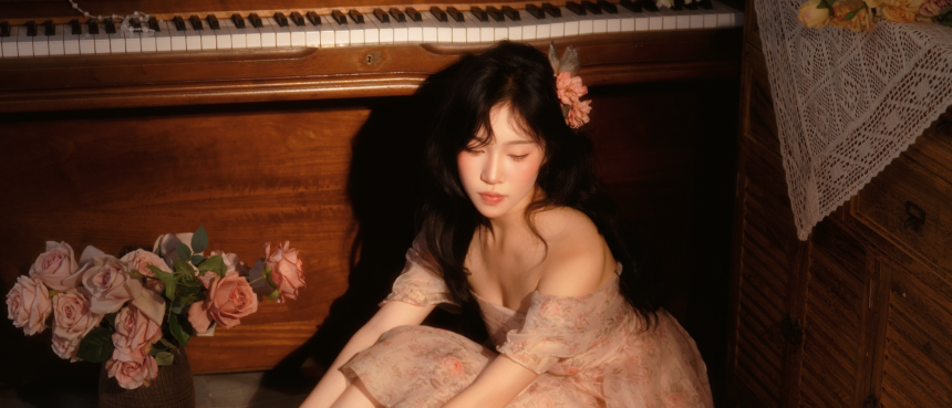 Clarity Liao sits beside a wooden piano in a soft floral off-the-shoulder dress with a flower in her hair, eyes closed in a warm, intimate setting with pink roses, promoting her single 'Have You.'