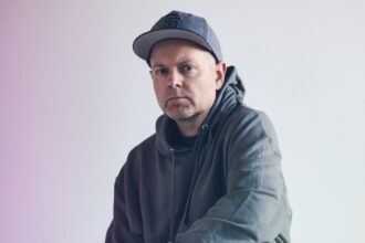 DJ Shadow, reissuing 'Endtroducing' for its 30th anniversary, poses in a hoodie and cap against a soft gradient backdrop.