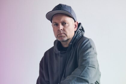 DJ Shadow, reissuing 'Endtroducing' for its 30th anniversary, poses in a hoodie and cap against a soft gradient backdrop.