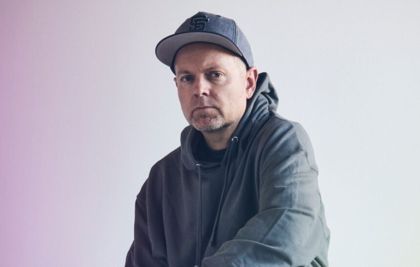 DJ Shadow, reissuing 'Endtroducing' for its 30th anniversary, poses in a hoodie and cap against a soft gradient backdrop.