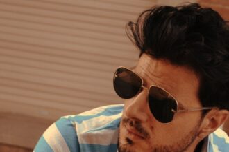 DJ Sparadise poses in aviator sunglasses and a blue-and-white striped polo shirt against a warm-toned backdrop, in press art for his single 'See U Again.'