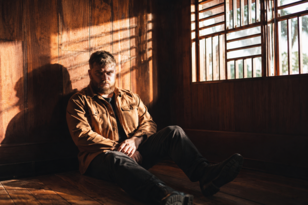 Irish singer Dave Lofts, whose single 'I'm All Yours' closes his debut EP 'Holy,' sits in a sunlit wood-paneled room in a moody portrait.