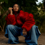 Dawn Richard crouches on a dirt path in a red fur coat, wide-leg jeans, and gold jewelry against lush greenery, in press art for her single 'chuuuuurch' and album 'Creole Culture.'