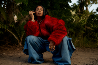 Dawn Richard crouches on a dirt path in a red fur coat, wide-leg jeans, and gold jewelry against lush greenery, in press art for her single 'chuuuuurch' and album 'Creole Culture.'