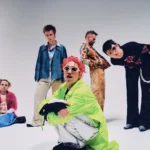 The five members of Fontaines D.C. pose in colorful, eclectic outfits against a plain grey backdrop, in press art for their 2026 arena tour and album 'Dopamine Chamber.'