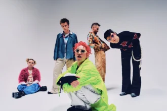 The five members of Fontaines D.C. pose in colorful, eclectic outfits against a plain grey backdrop, in press art for their 2026 arena tour and album 'Dopamine Chamber.'