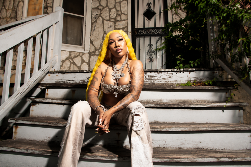 Glizzy Glow sits on a worn front stoop with bright yellow hair, tattoos, and layered jewelry, in press art for her debut mixtape 'Trench Baddie.'