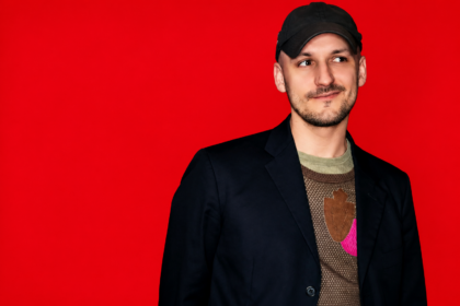 Jacques Greene, announcing his club album 'jg,' poses in a black blazer and cap against a bright red backdrop.