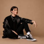 Press-style image for a Jade LeMac feature, showing a young woman in a black leather jacket and Converse high-tops seated against a tan backdrop.