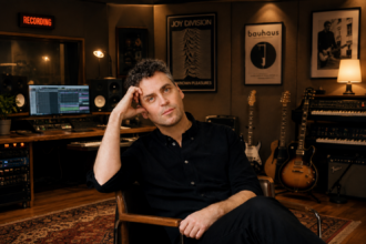 ames Ellis Ford sits in a warmly lit recording studio dressed in black, surrounded by guitars, synths, and framed Joy Division and Bauhaus posters, promoting his second solo album 'Lost In Another World.'