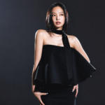 BLACKPINK's Jennie poses in an elegant black one-shoulder dress against a dark backdrop in a photo shoot, around the announcement of her solo EP 'Fallen Angel.'