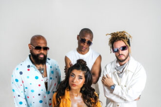 The four members of Latin collective Vale! ULB pose against a plain light backdrop in colorful, stylish outfits, with vocalist Val at the front, promoting their single 'Devórame.'