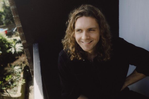 Lewis Knaggs smiles in a black shirt, seated in a doorway with greenery behind him, in press art for his single 'Forget How to Forgive.'