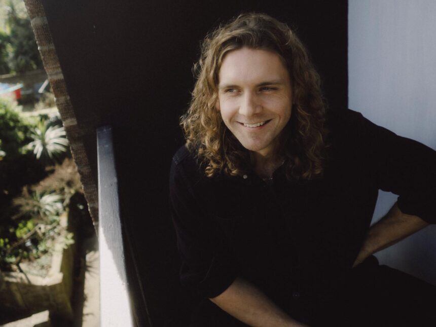 Lewis Knaggs smiles in a black shirt, seated in a doorway with greenery behind him, in press art for his single 'Forget How to Forgive.'