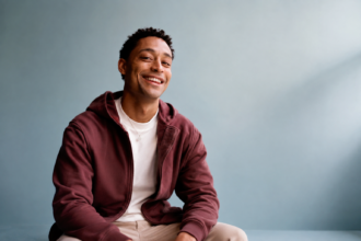 Loyle Carner smiles in a maroon hoodie against a grey backdrop, in press-style art tied to his Reading 2026 set and his comments on J.K. Rowling and toxic masculinity.