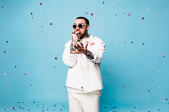 The late Mac Miller poses in a white jacket and sunglasses against a blue backdrop with falling petals, marking the 10th-anniversary reissue of 'The Divine Feminine.'