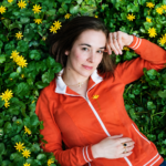 Margaret Glaspy lies among lush green foliage and bright yellow flowers, wearing an orange zip-up jacket and looking calmly toward the camera in a natural outdoor portrait