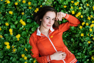 Margaret Glaspy lies among lush green foliage and bright yellow flowers, wearing an orange zip-up jacket and looking calmly toward the camera in a natural outdoor portrait