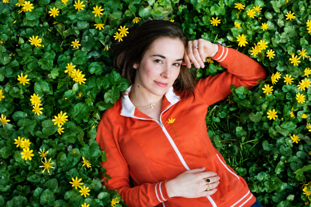 Margaret Glaspy lies among lush green foliage and bright yellow flowers, wearing an orange zip-up jacket and looking calmly toward the camera in a natural outdoor portrait