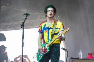 Modest Mouse live photo tied to the Simon O'Connor tour news, a band guitarist performing in a "VOTE" shirt with a green guitar.
